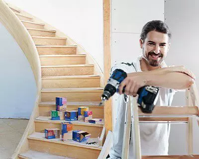 Mann mit Akkubohrer vor einer Holztreppe mit diversen Schraubenverpackungen